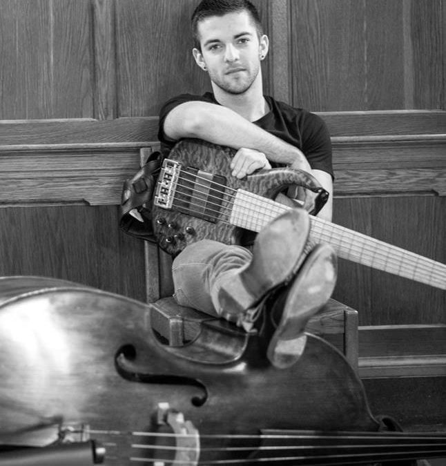Dr. Andrew O'Connor black white photo. He is seated with his feet propped up on a double bass turned sideways and an electric bass crossed over his lap, with his arm resting on top.