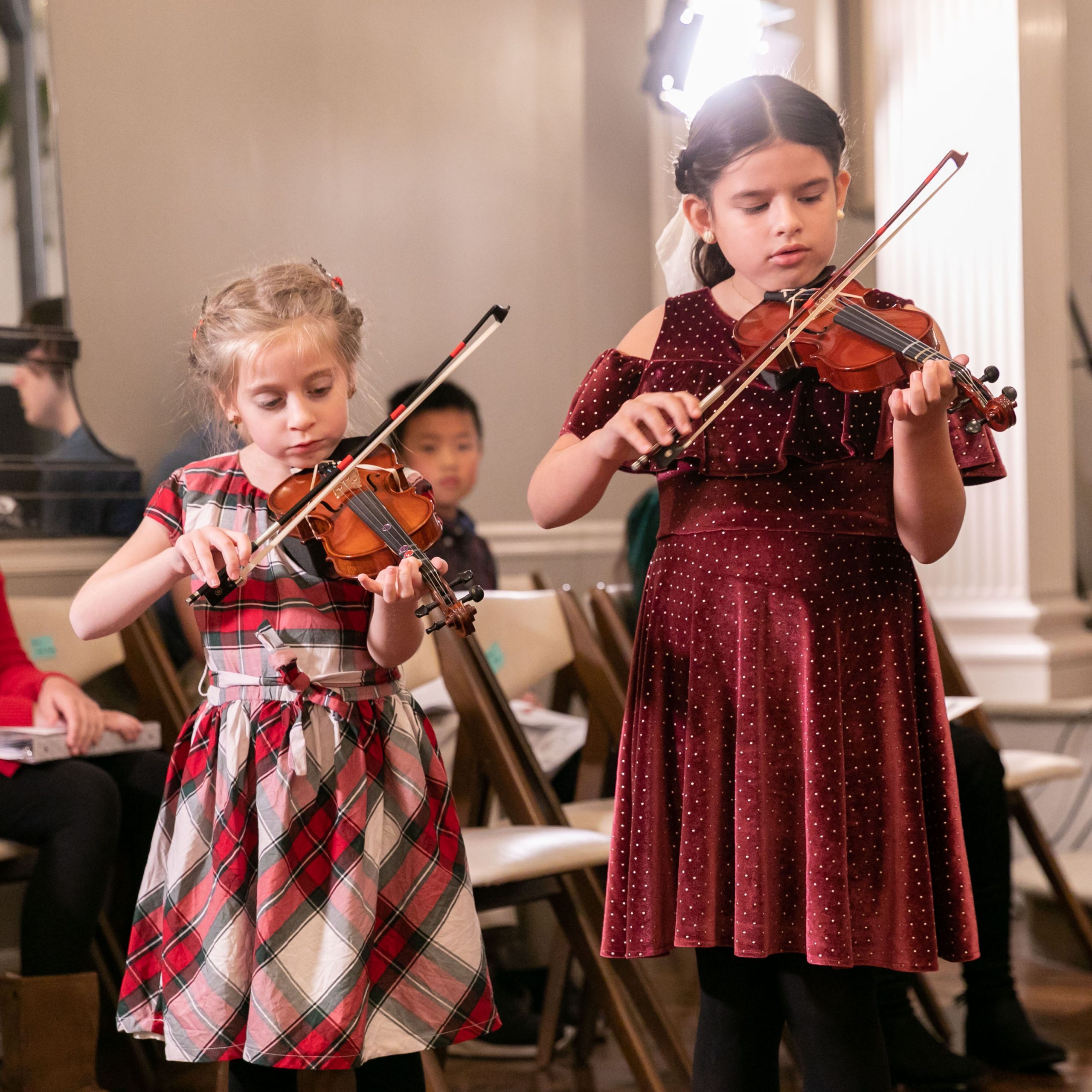 Two young female violin students in festive holiday dresses playing a violin duet in an elegant recital hall at Community Music School Allentown CMS