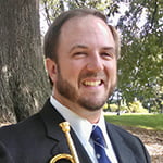 Chris Klump candid headshot in a suit and tie, holding a trumpet with a background of trees.