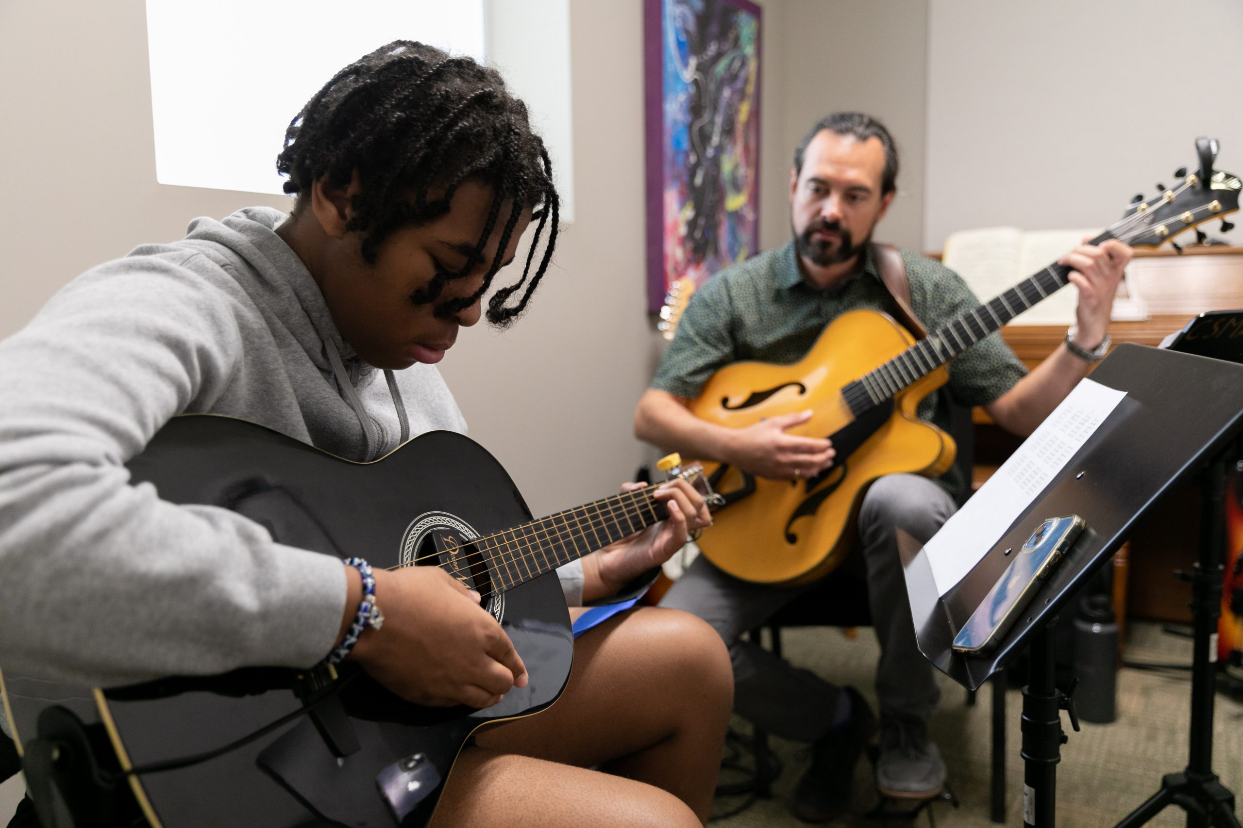 A private guitar lesson at Community Music School in the Martin Guitar Studio.  On the right, teacher Joe Wagner with a blue button down shirt, dark hair pulled back in a ponytail, and holding a white, black, and brown electric guitar, points to music on a stand.  Teenage student, Andrew, wearing a blue polo shirt, and holding a black and white electric guitar looks on intensely. 
