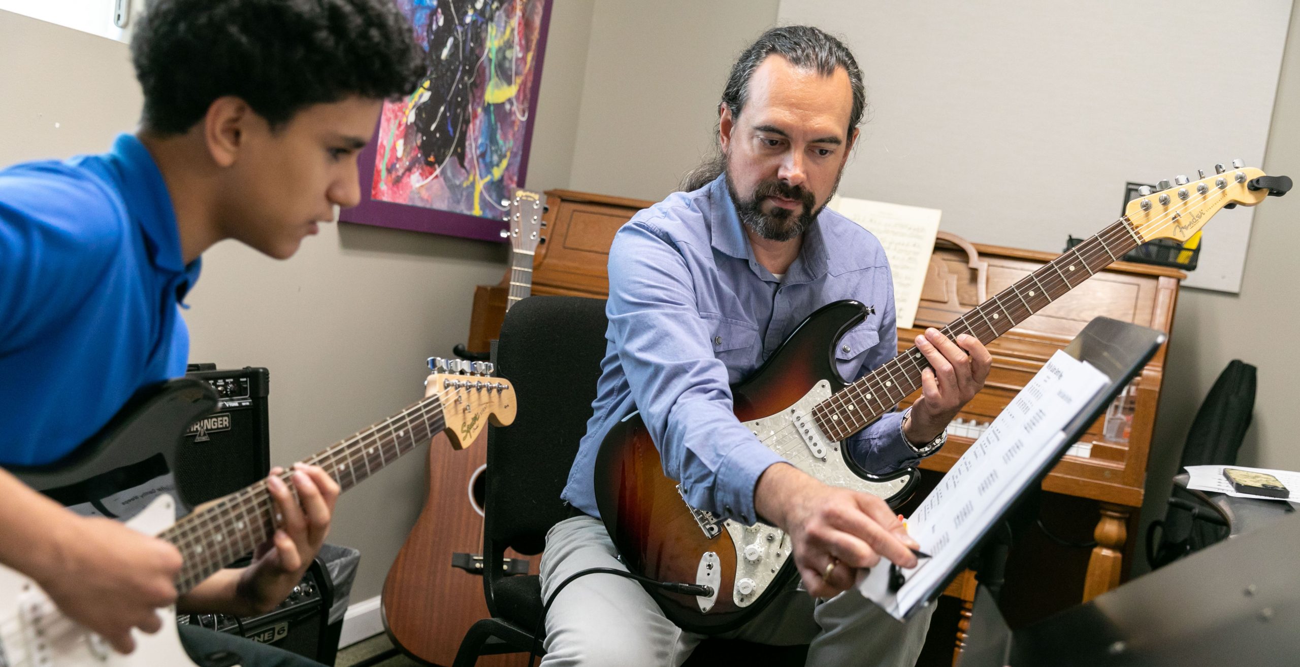 A private guitar lesson at Community Music School in the Martin Guitar Studio. On the right, teacher Joe Wagner with a blue button down shirt, dark hair pulled back in a ponytail, and holding a white, black, and brown electric guitar, points to music on a stand. Teenage student, Andrew, wearing a blue polo shirt, and holding a black and white electric guitar looks on intensely.